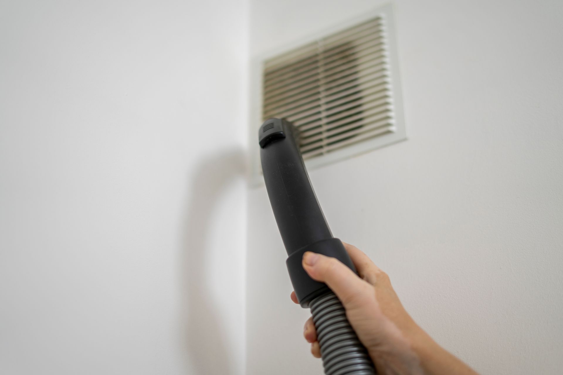 A person uses a vacuum cleaner nozzle to remove dust from a white wall-mounted air vent.