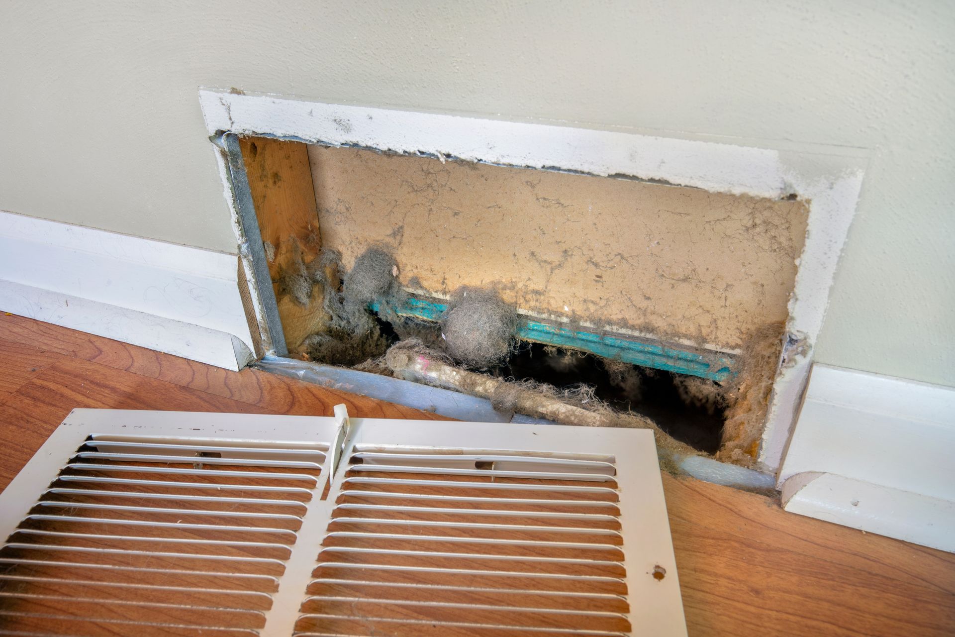 A detached metal air vent cover sits on a floor in front of a wall cavity filled with accumulated dust and debris.