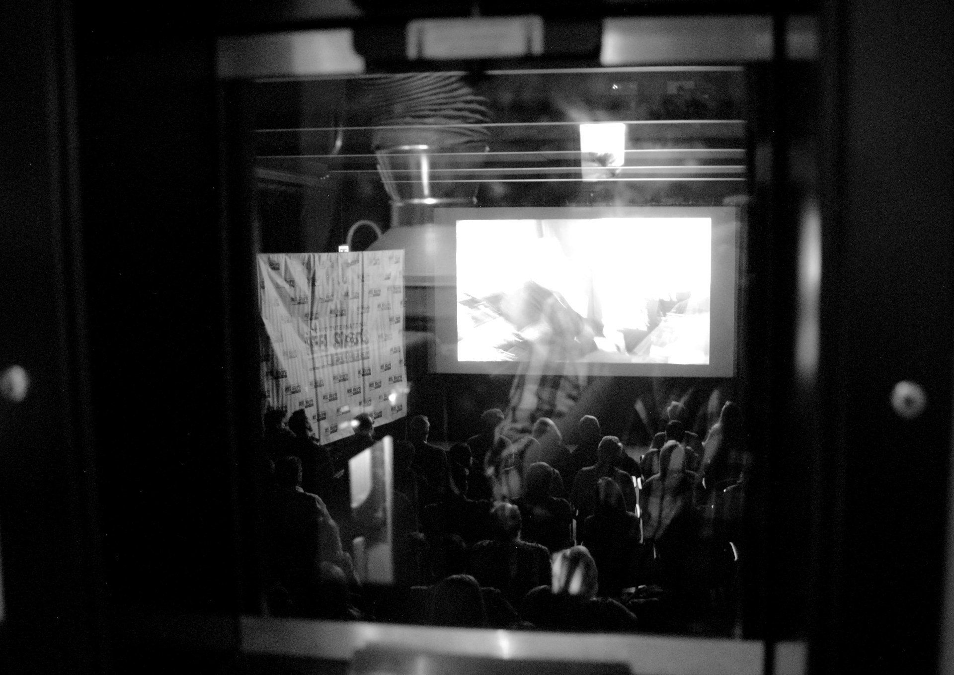 Audience watching a bright screen, viewed through a dark doorway. Includes a podium, projector, and bright light.