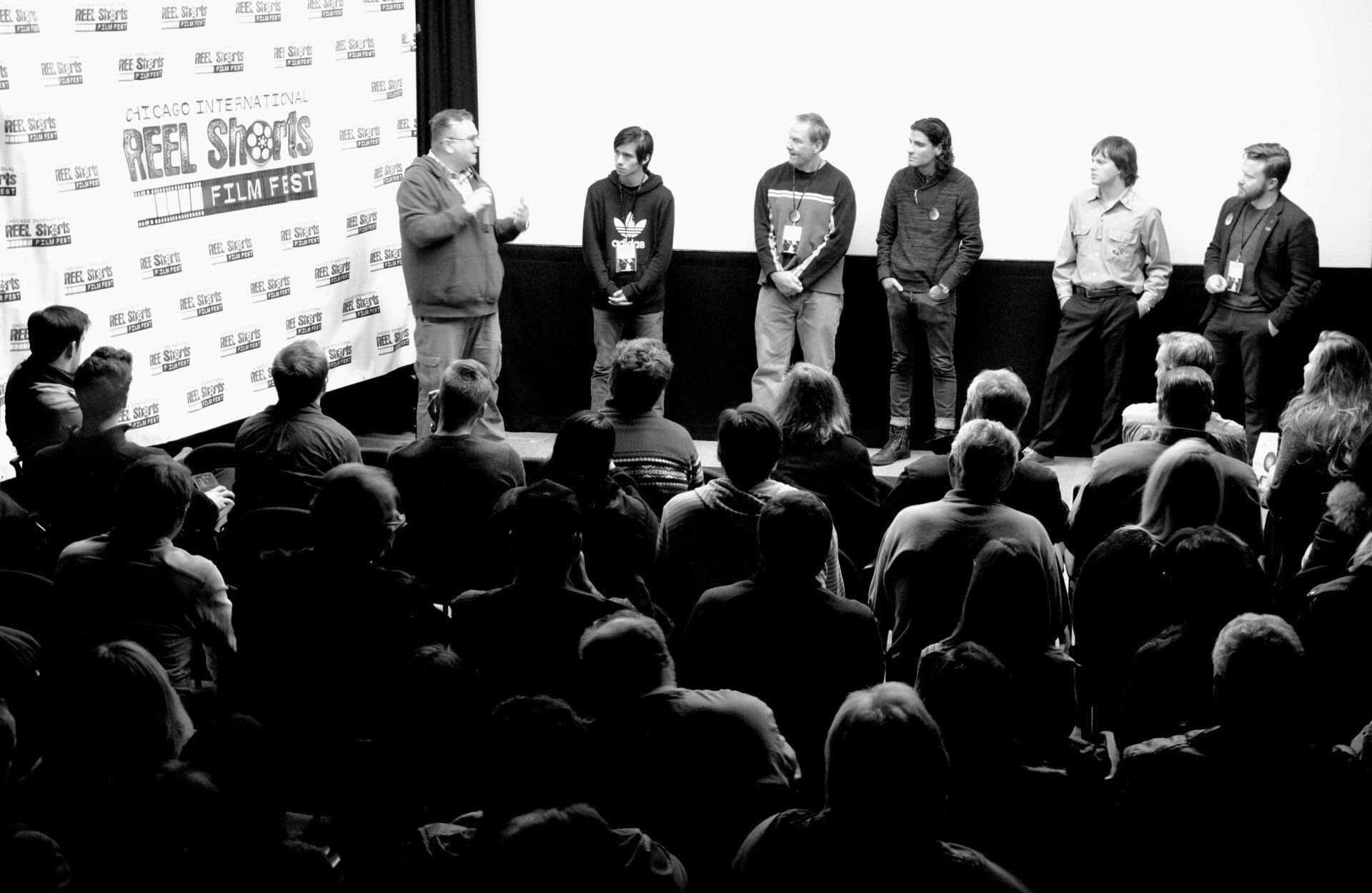 A panel of people standing on a stage speaking to an audience. Black and white.