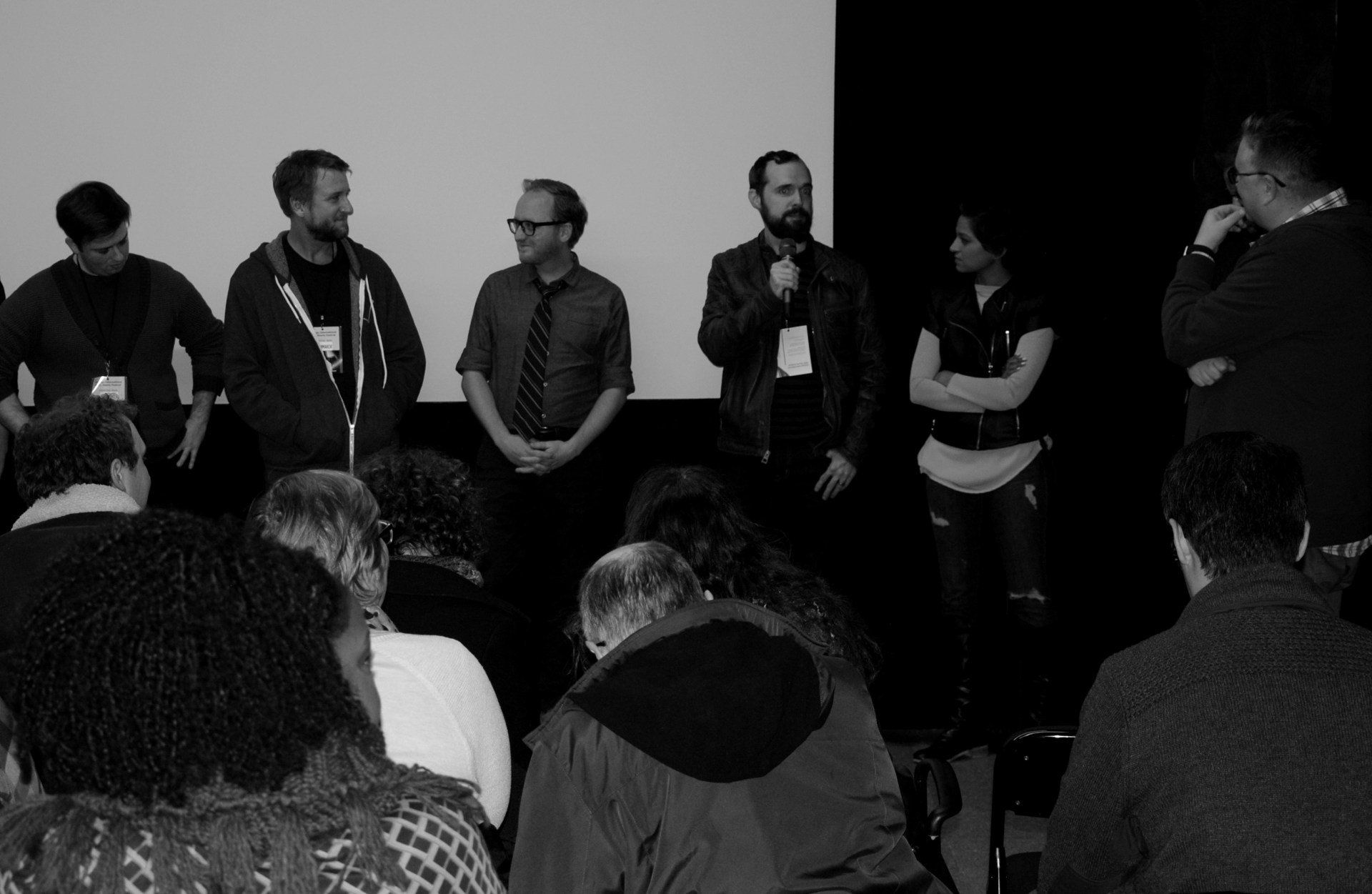 Panel of six people on stage, addressing an audience in a dimly lit room.