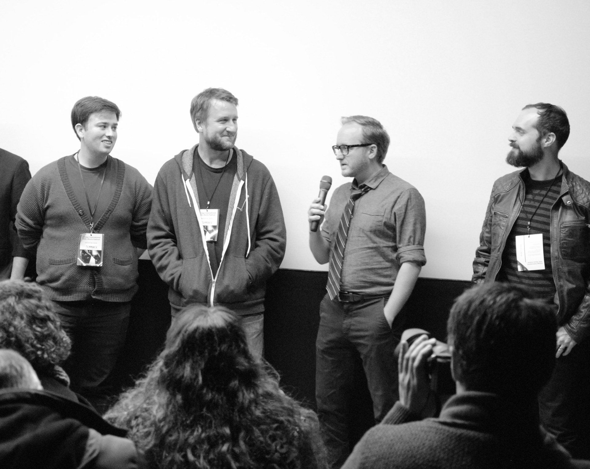 Group of men on stage, one with a microphone, facing audience.