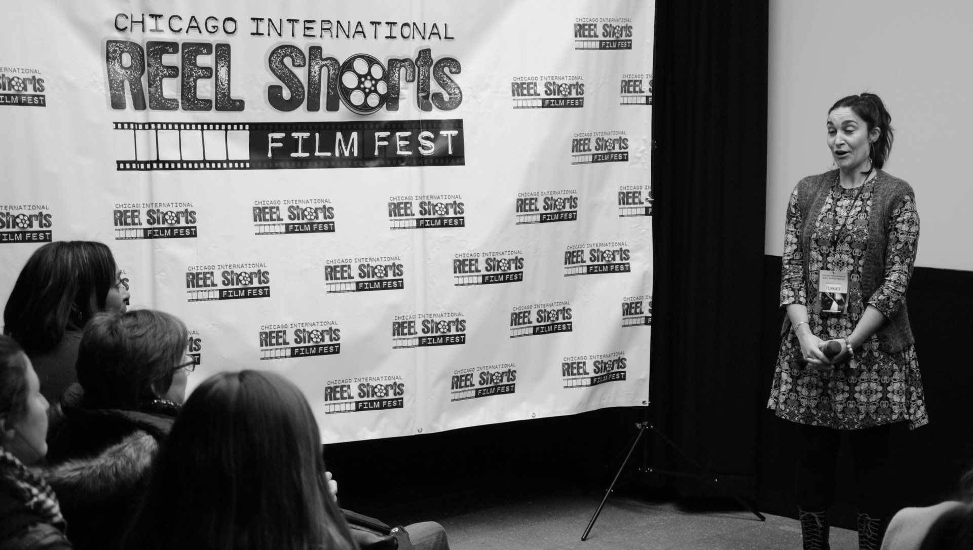 Woman at a film festival speaks to an audience. Large banner behind. Black and white photo.