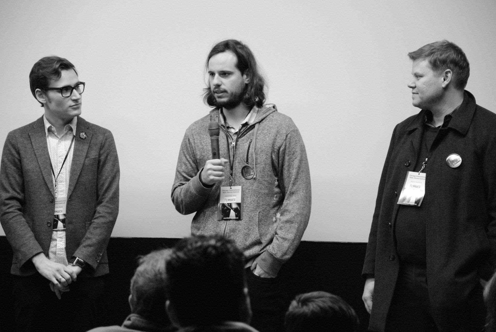 Three men on stage: one speaking into a mic, flanked by two others. Black and white, crowd in foreground.