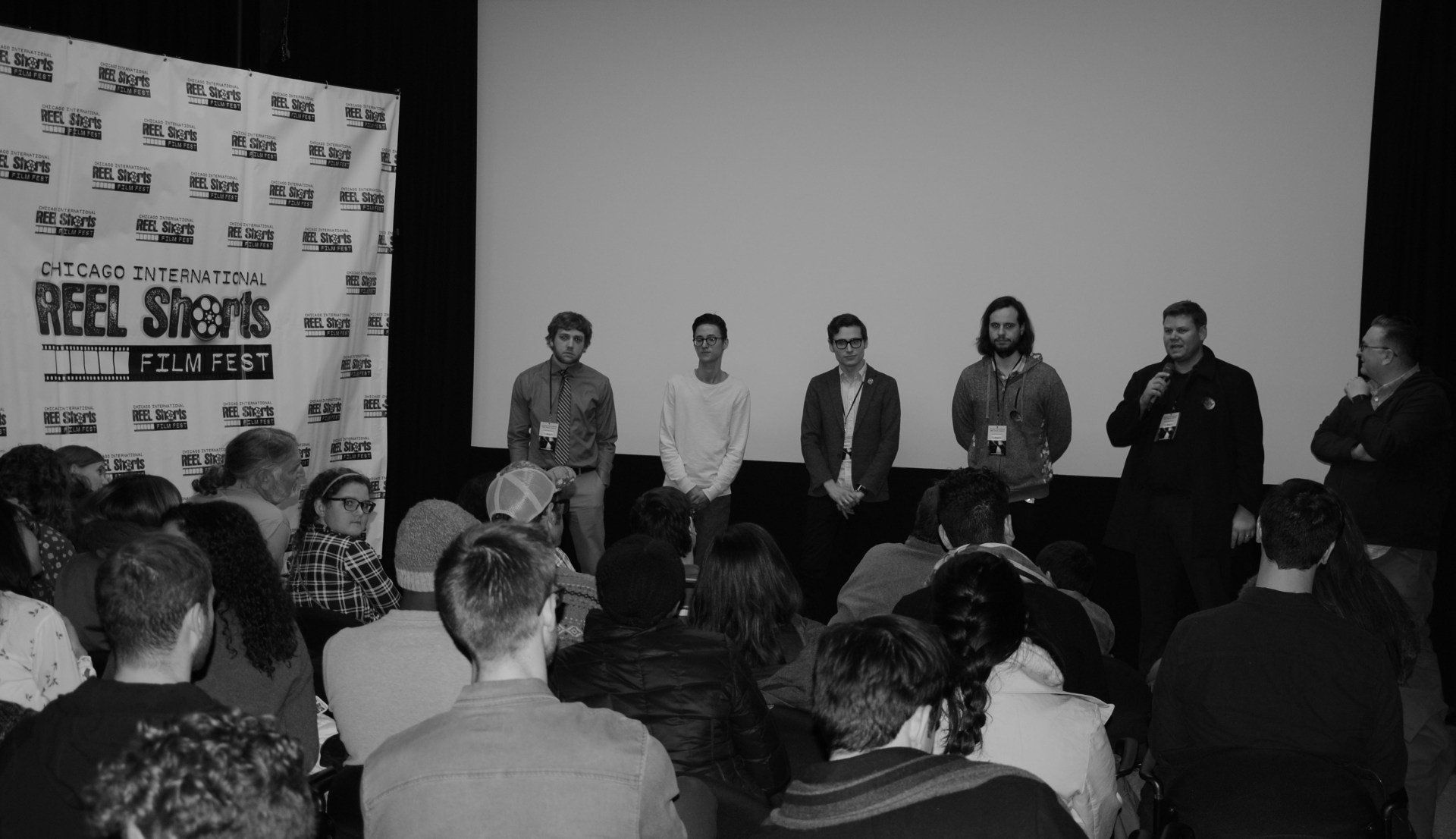 Panel of filmmakers on stage at a film festival, addressing an audience. Black and white.