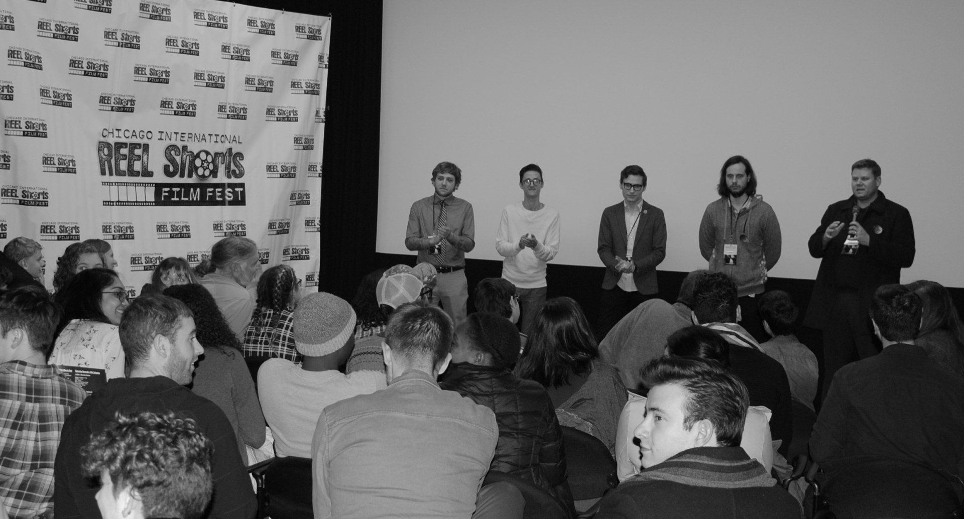 Panel discussion at a film festival. Five men on stage, audience seated in front. Black and white photo.