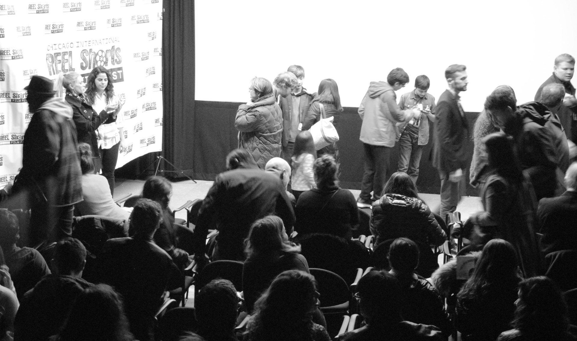 Audience watches a film screening. People on stage, spotlight on screen, black and white photo.