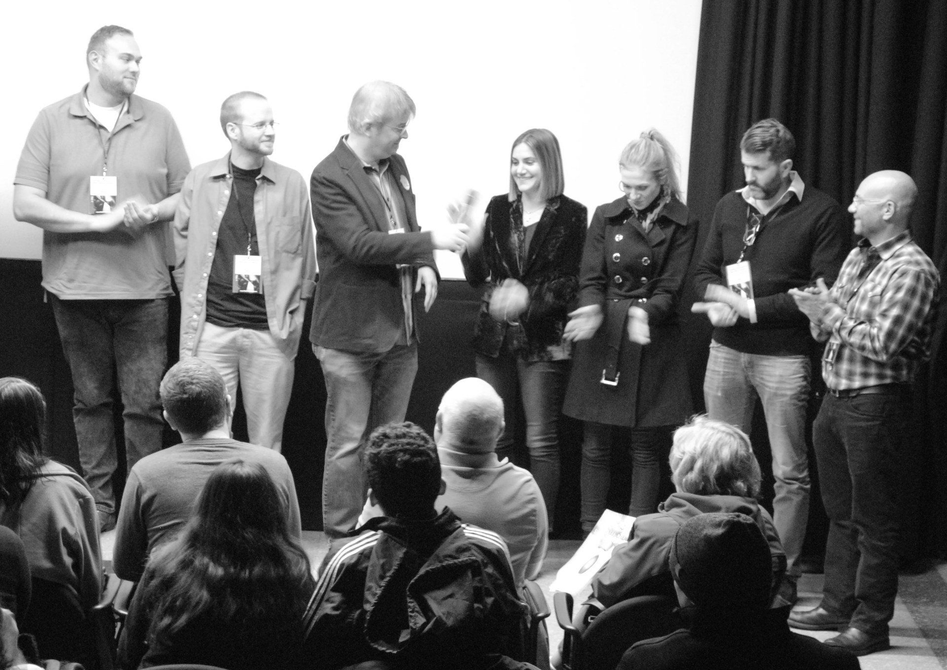 Group of people on stage, facing an audience. Some are applauding, some are smiling, and some are wearing lanyards.