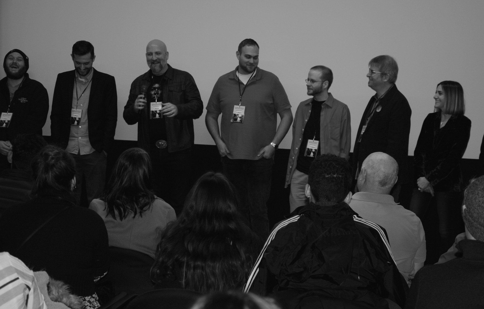 Panel of seven people on a stage facing an audience. Black and white photo.