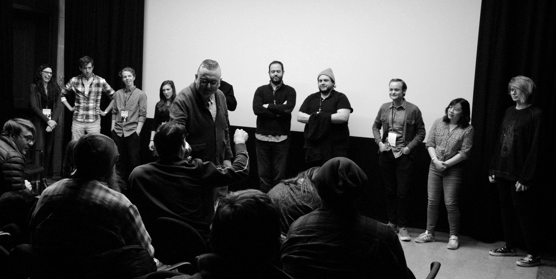 A group of people onstage in a theater, facing an audience. Black and white.