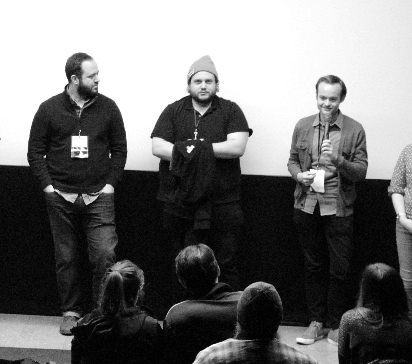 Three men stand on a stage, facing an audience, in front of a white screen. One holds a microphone, speaking.