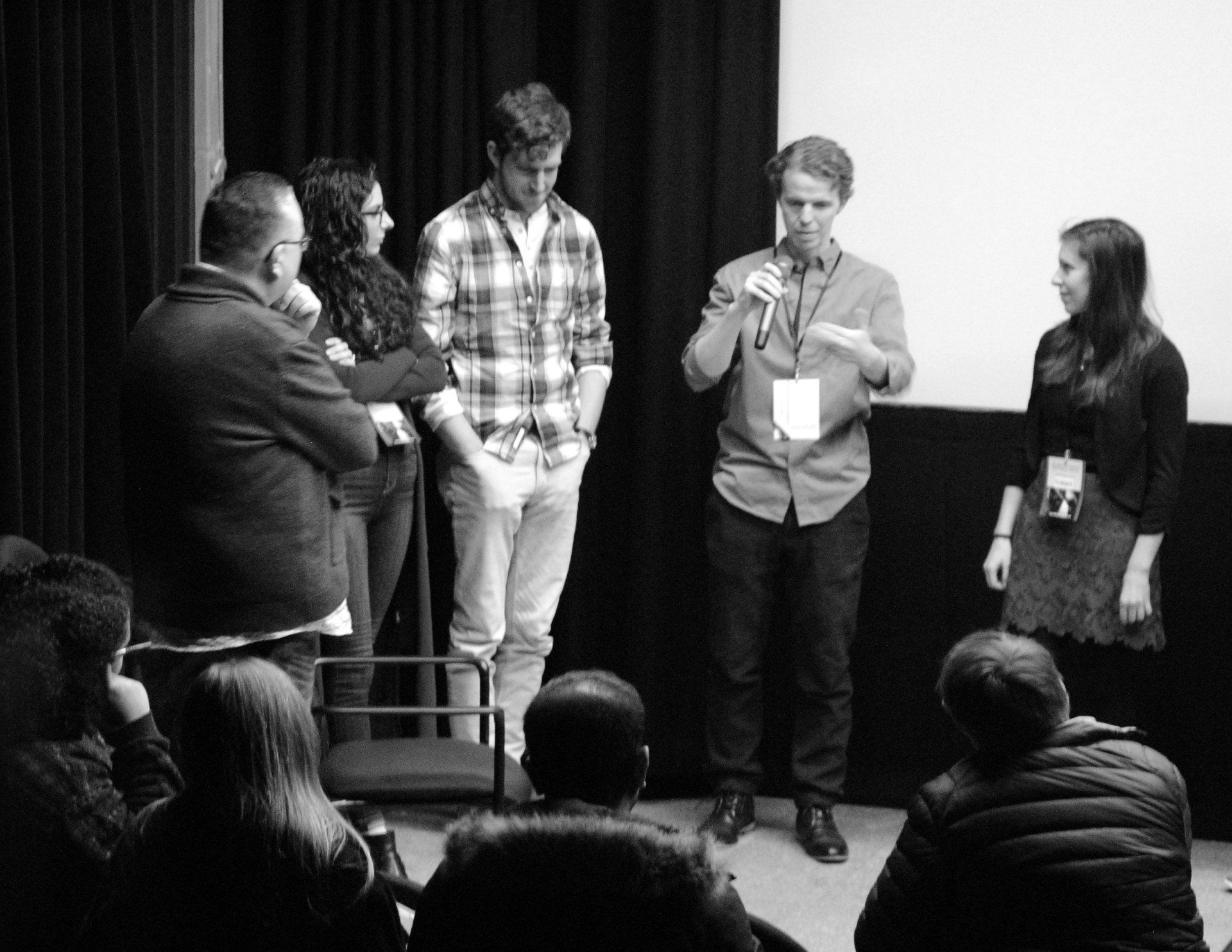 A group of people on a stage at a film screening. A man speaks into a microphone, others listen and watch.