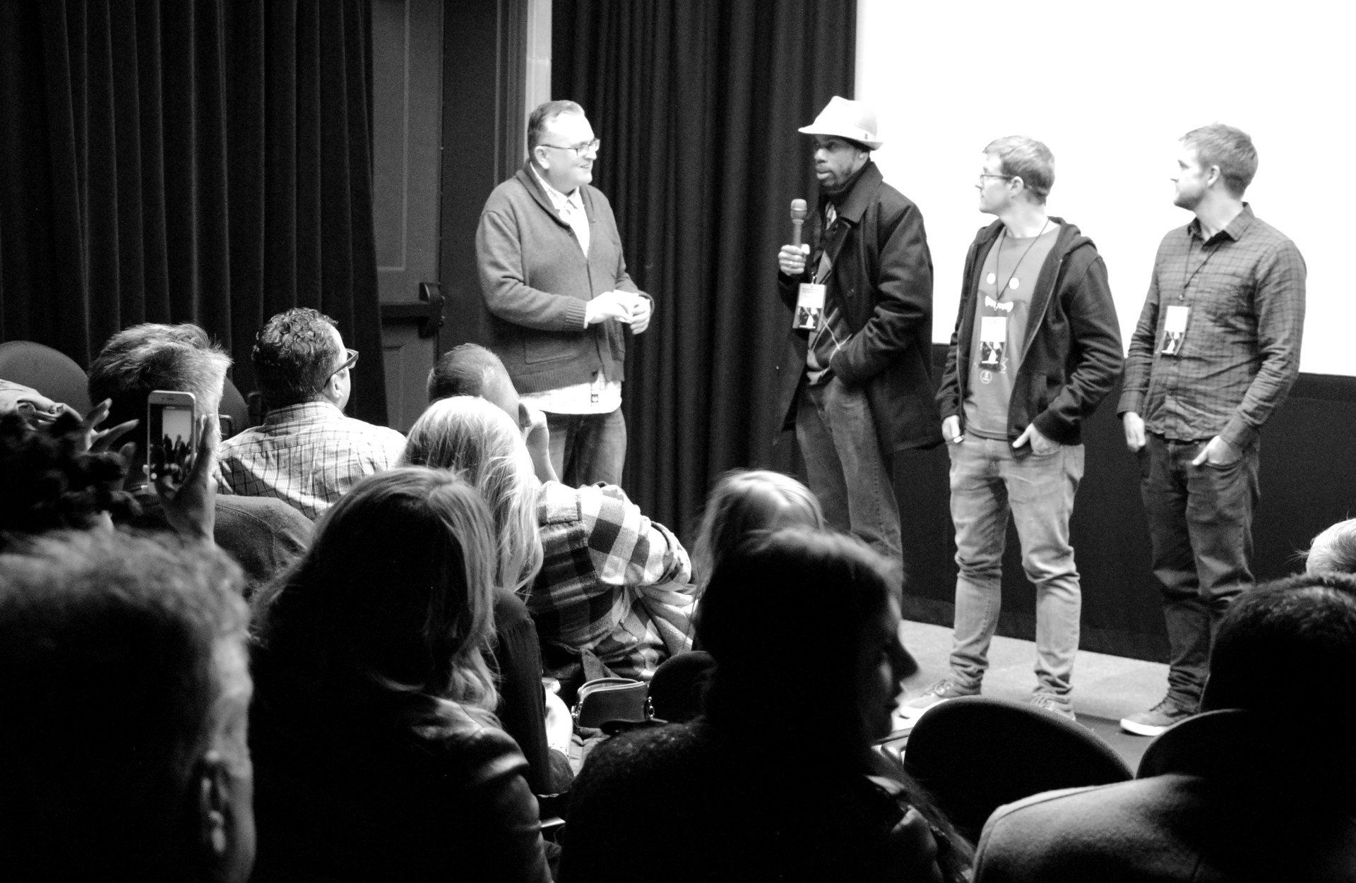 Audience watches panel discussion with four men on stage, one speaking into a microphone. Black and white.