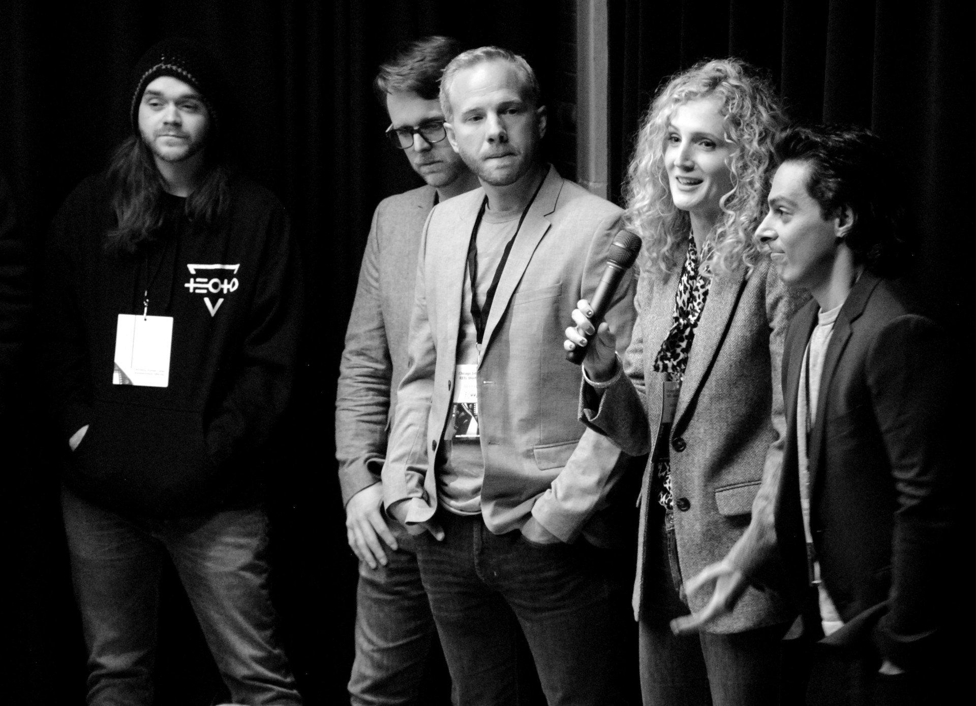 Group of five people on stage, a woman with a microphone speaks; black and white.