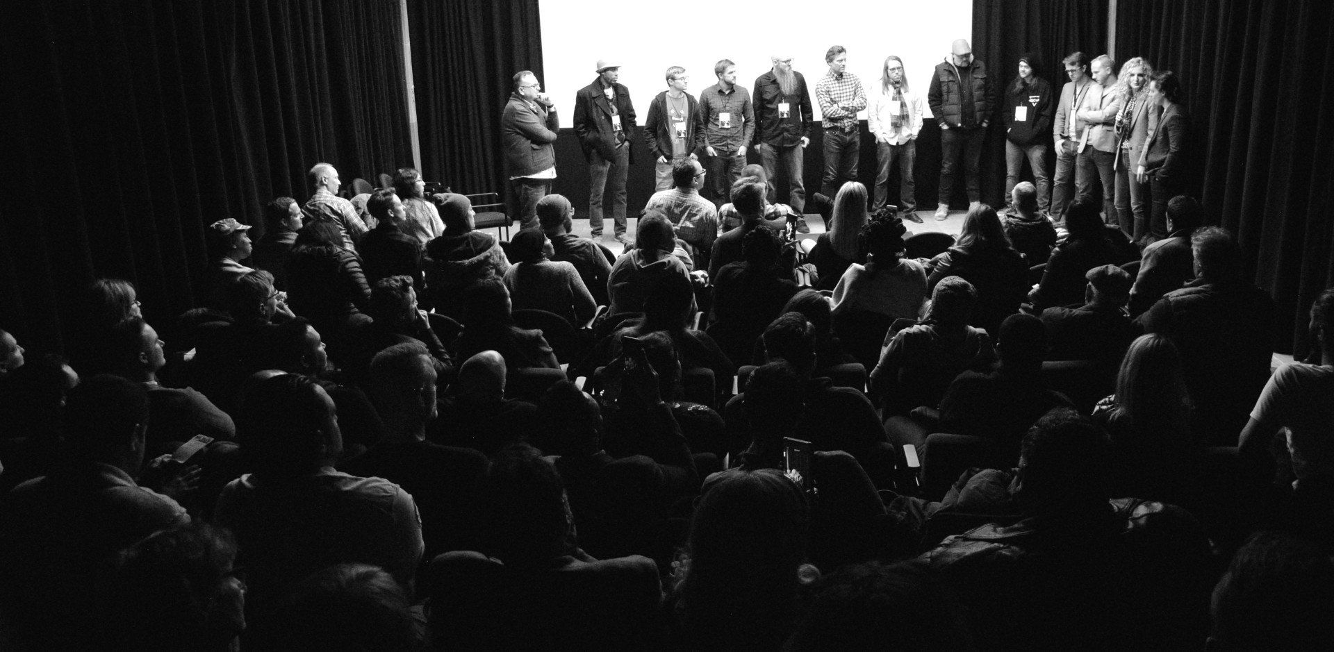 Audience seated in a dark auditorium facing a brightly lit stage with a group of people standing on it.