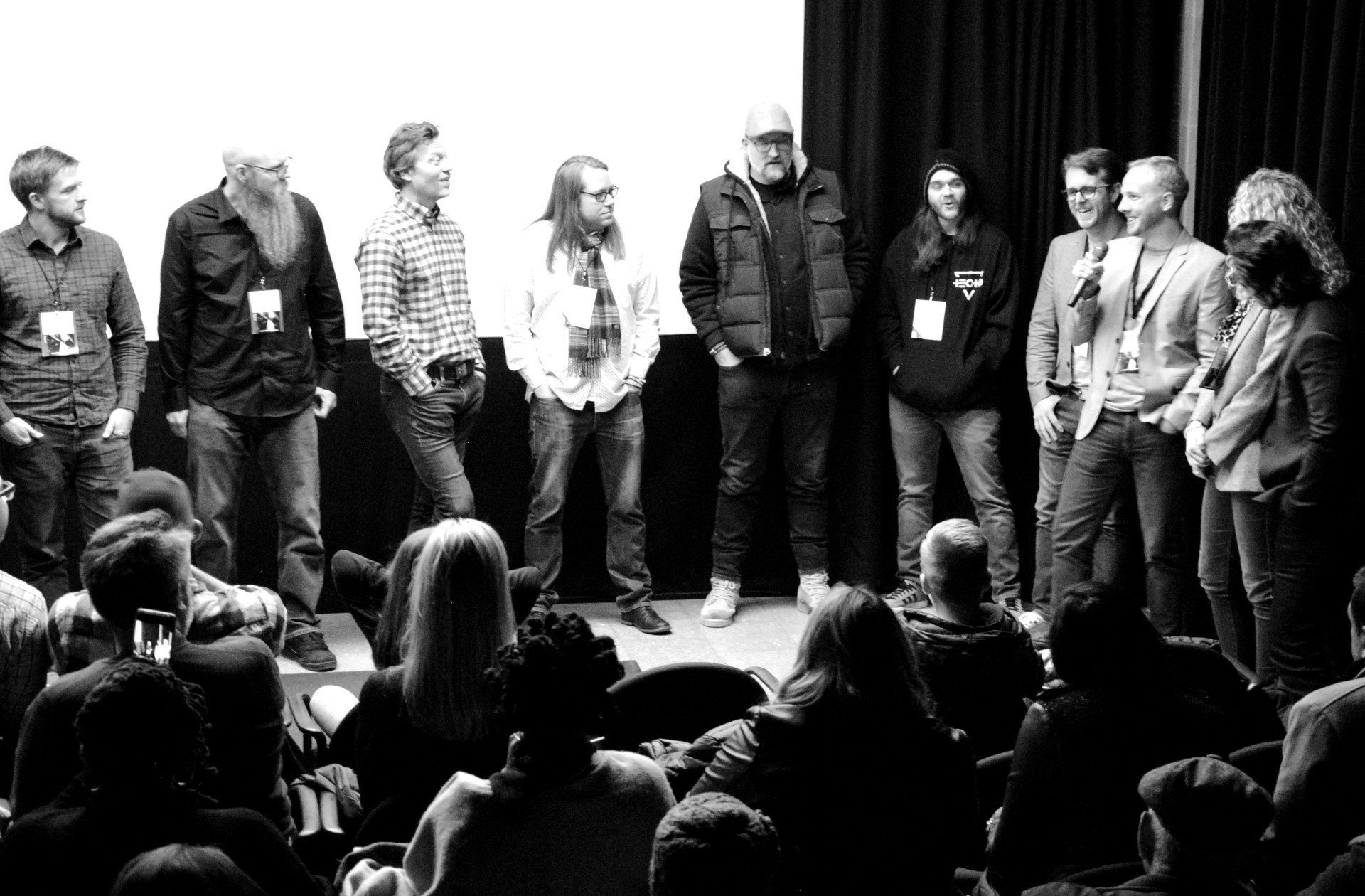 A group of people on a stage, facing an audience in a dark room. Black and white photo.