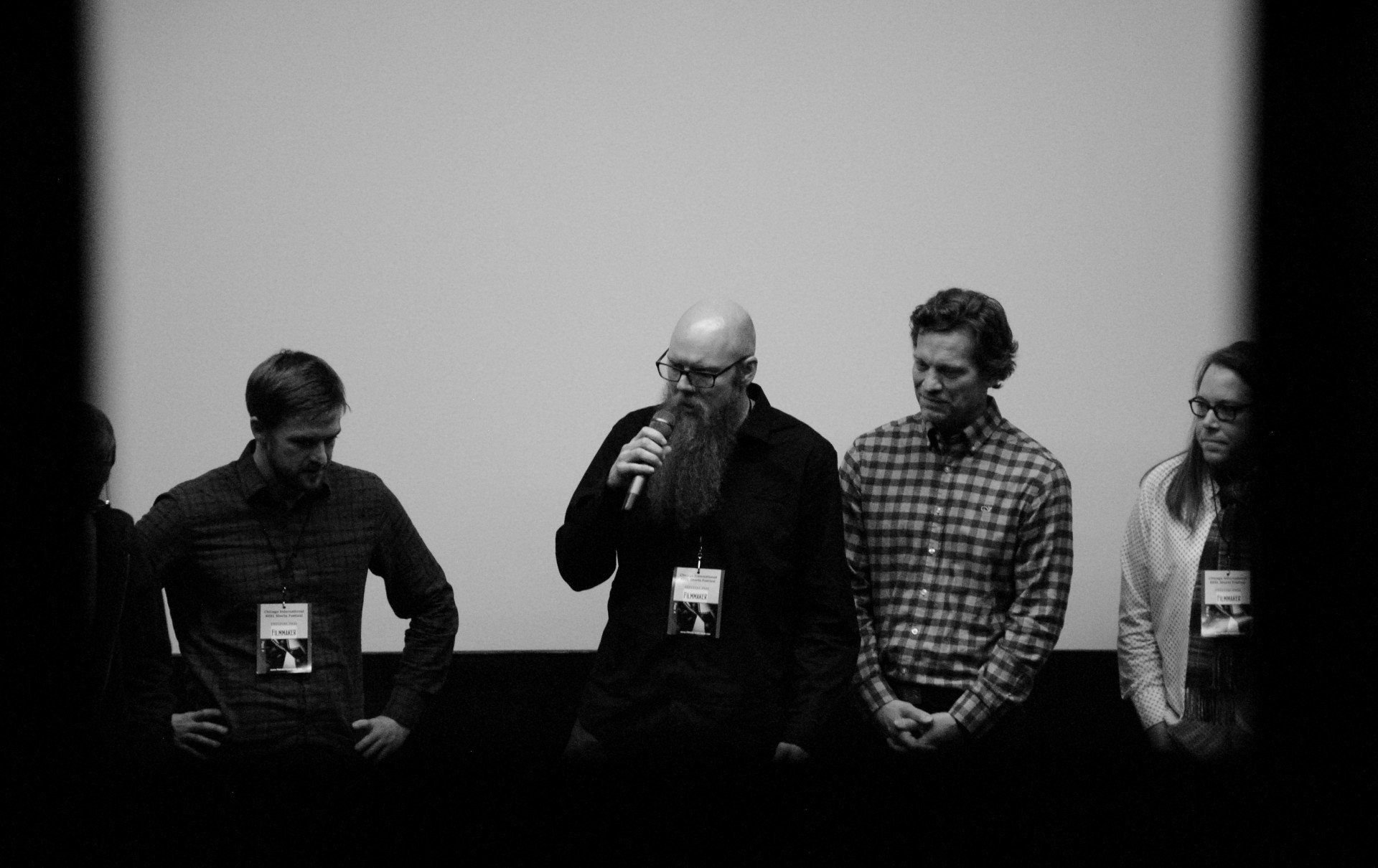 A panel of four people stands on a stage in front of a screen. One man speaks into a microphone.