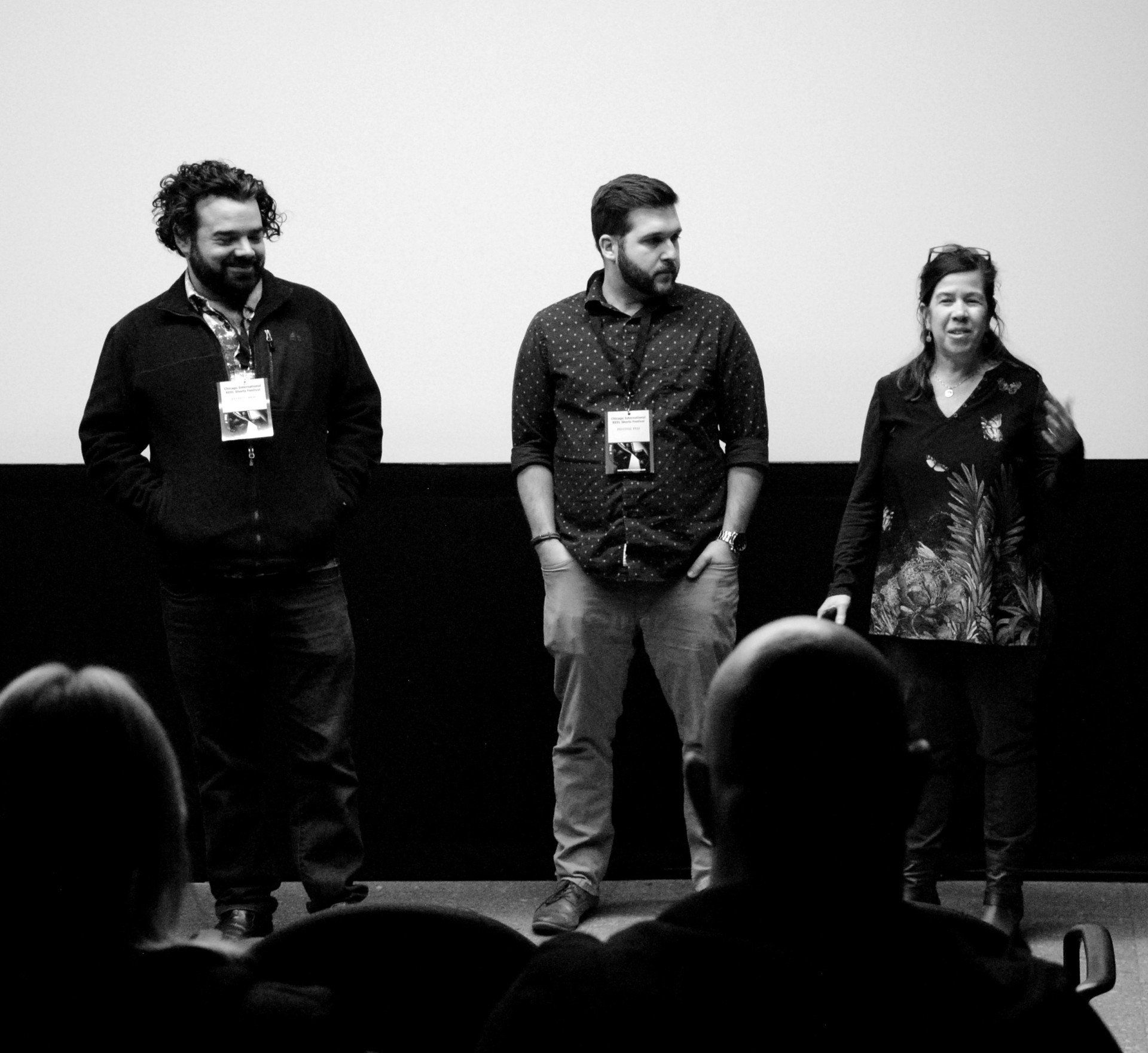 Three people stand onstage, facing an audience in a dark theater.