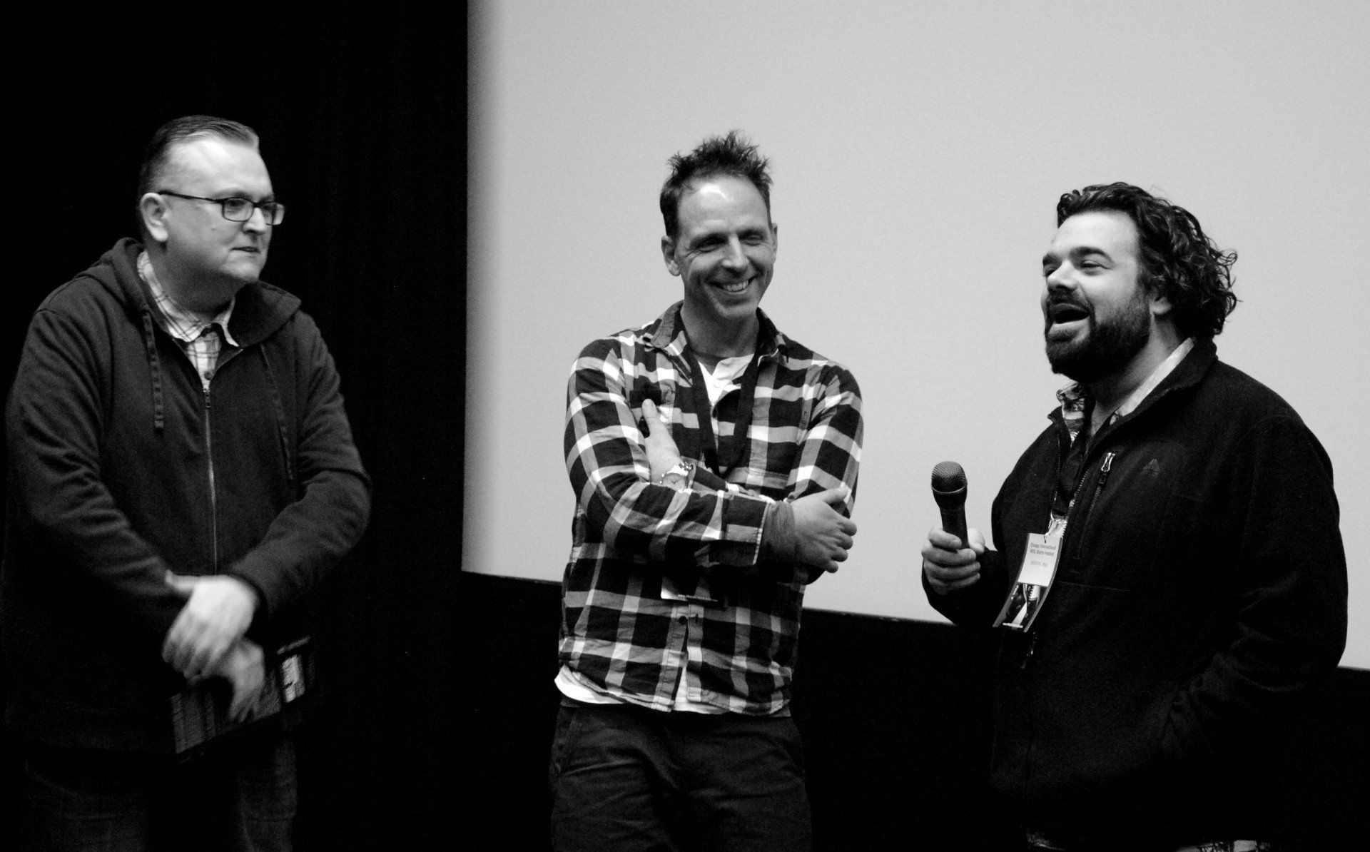Three men on stage, one holding a microphone. Two smile, the center one with crossed arms, a screen backdrop.