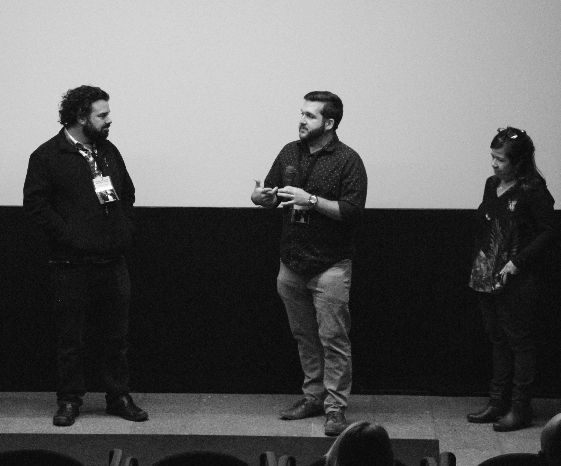 Three people on stage talking: a man gestures while speaking to the other two, in front of a plain white screen.