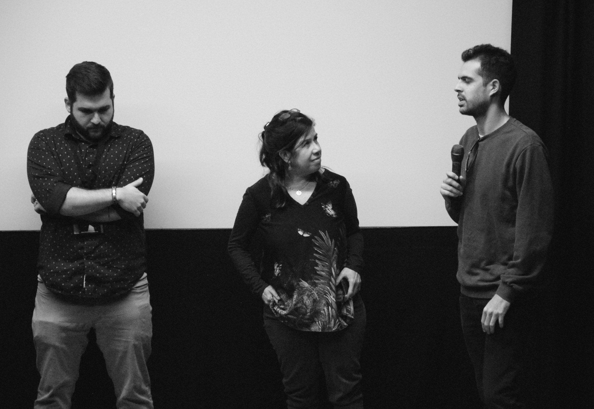 Three people stand on a stage. A man speaks into a microphone, another man has crossed arms, and a woman stands between them.