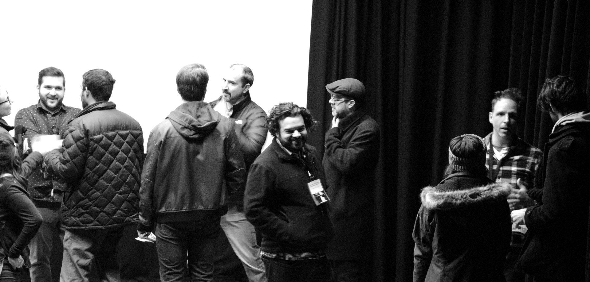 A group of people socializing indoors, some are facing each other, near a white wall and black curtain.