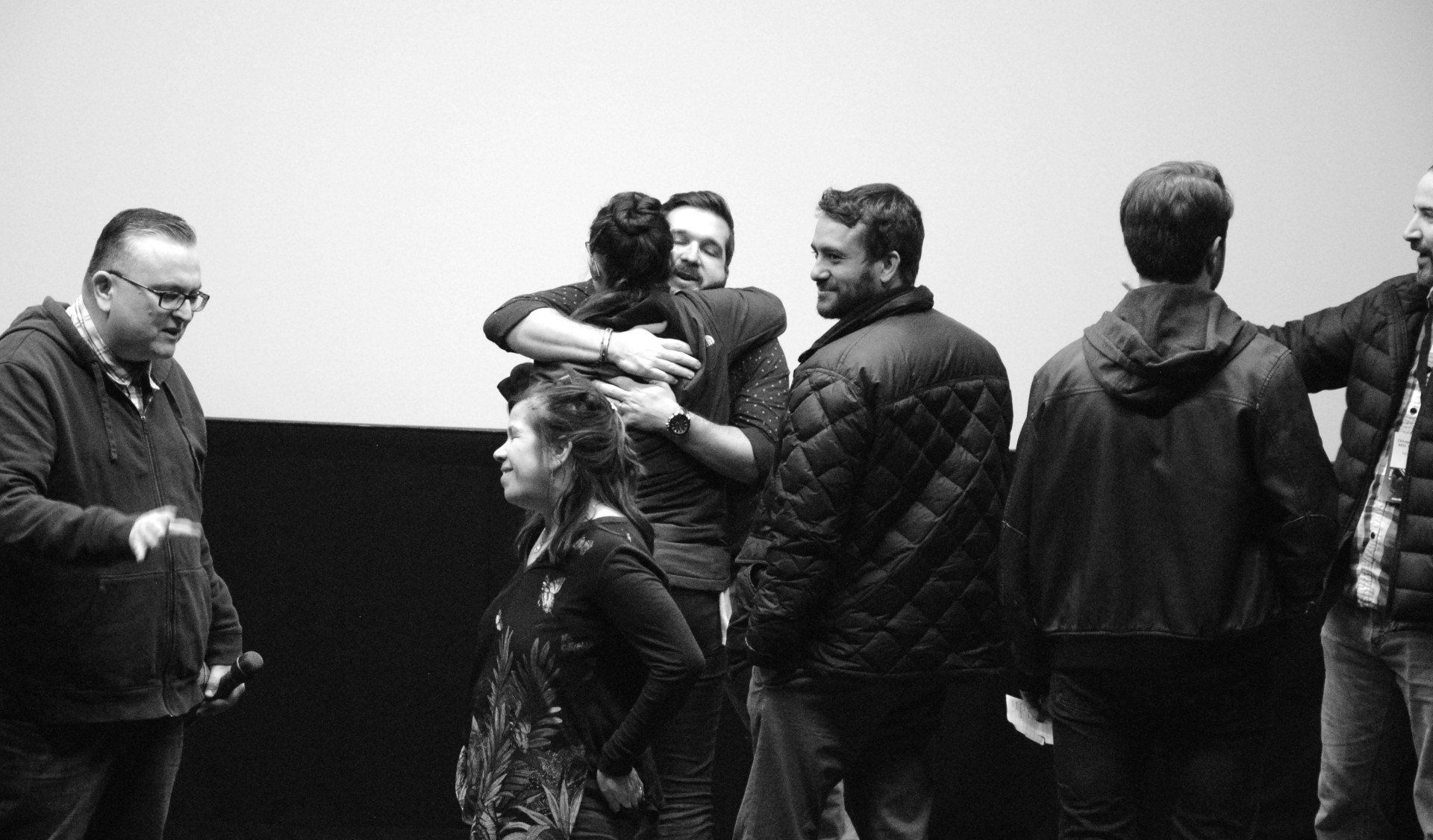 Group of people on stage, some hugging, in front of a screen. Black and white photo.