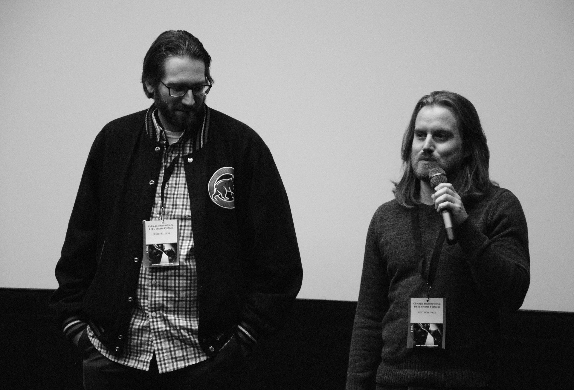 Two men, one with a microphone, stand on a stage in front of a screen. Both wear name tags and have beards.