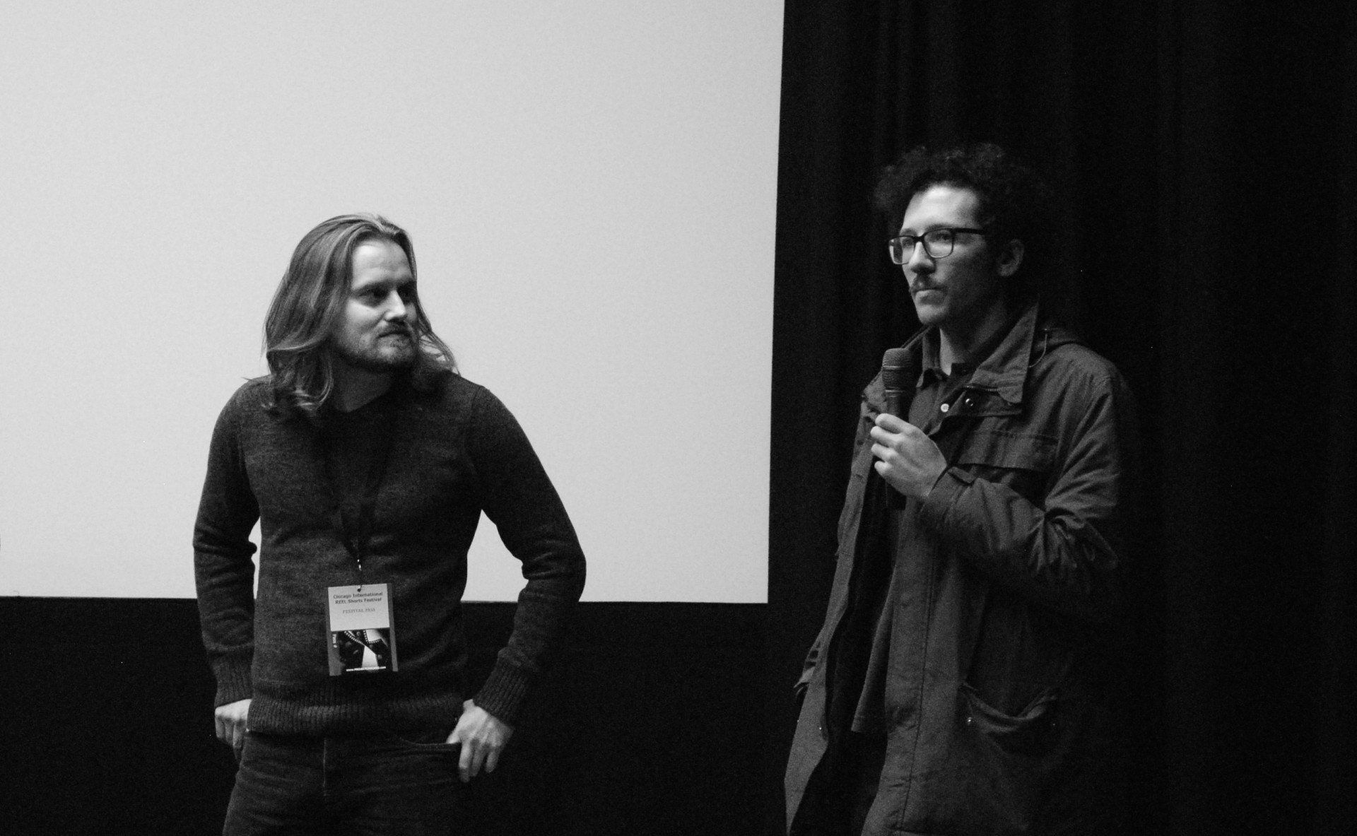 Two men stand by a screen. One holds a mic, speaking. Black and white photo.