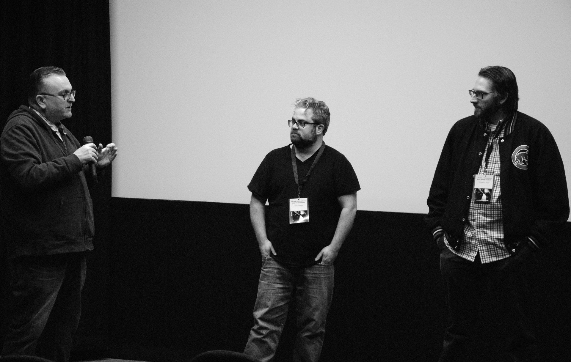 Three men on a stage in front of a screen. One speaks into a microphone, while the other two listen.