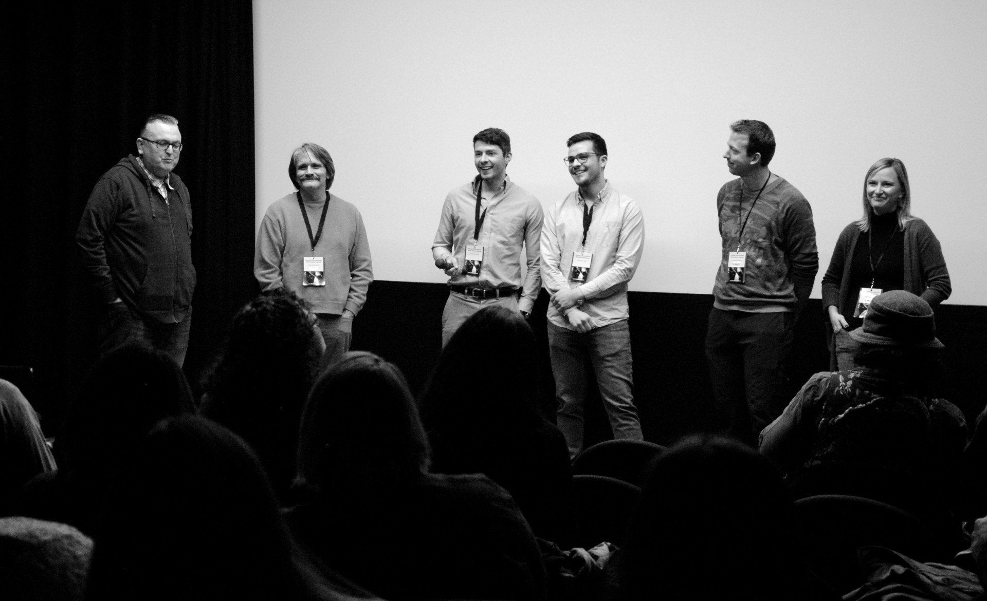 A group of six people on stage in front of a screen, facing a seated audience. Black and white photo.
