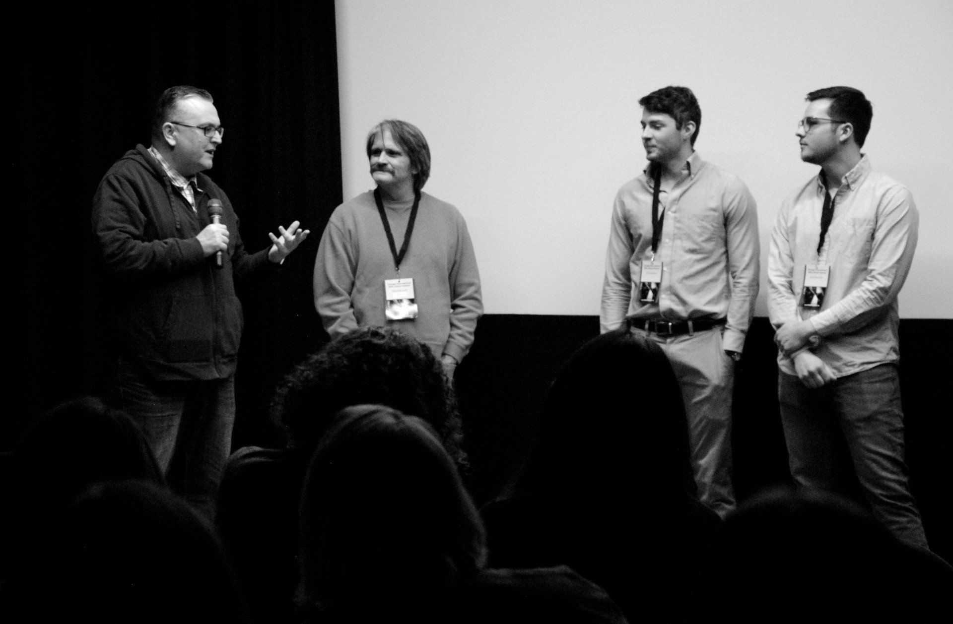 A man speaks into a microphone while three others stand on a stage in front of an audience.