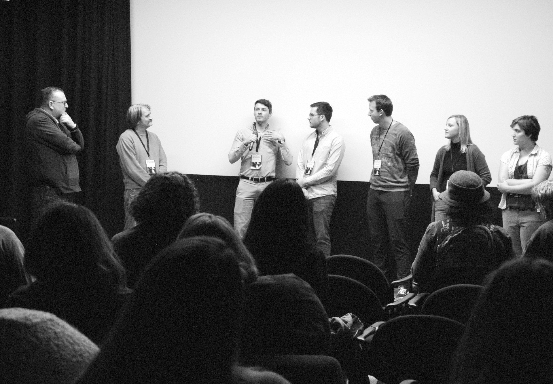 Panel of seven people speaking on stage in front of a movie screen. Audience members in chairs.
