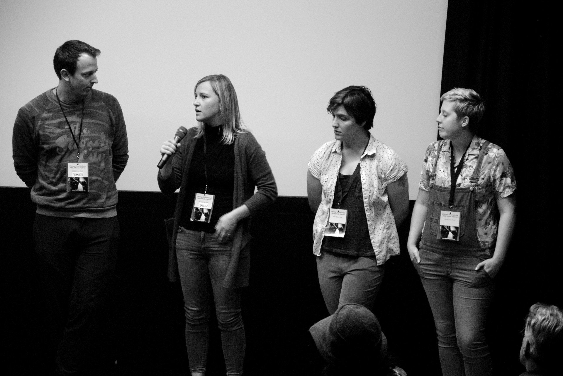 Panel of four people standing on stage, one with a microphone, in front of a screen. Black and white.