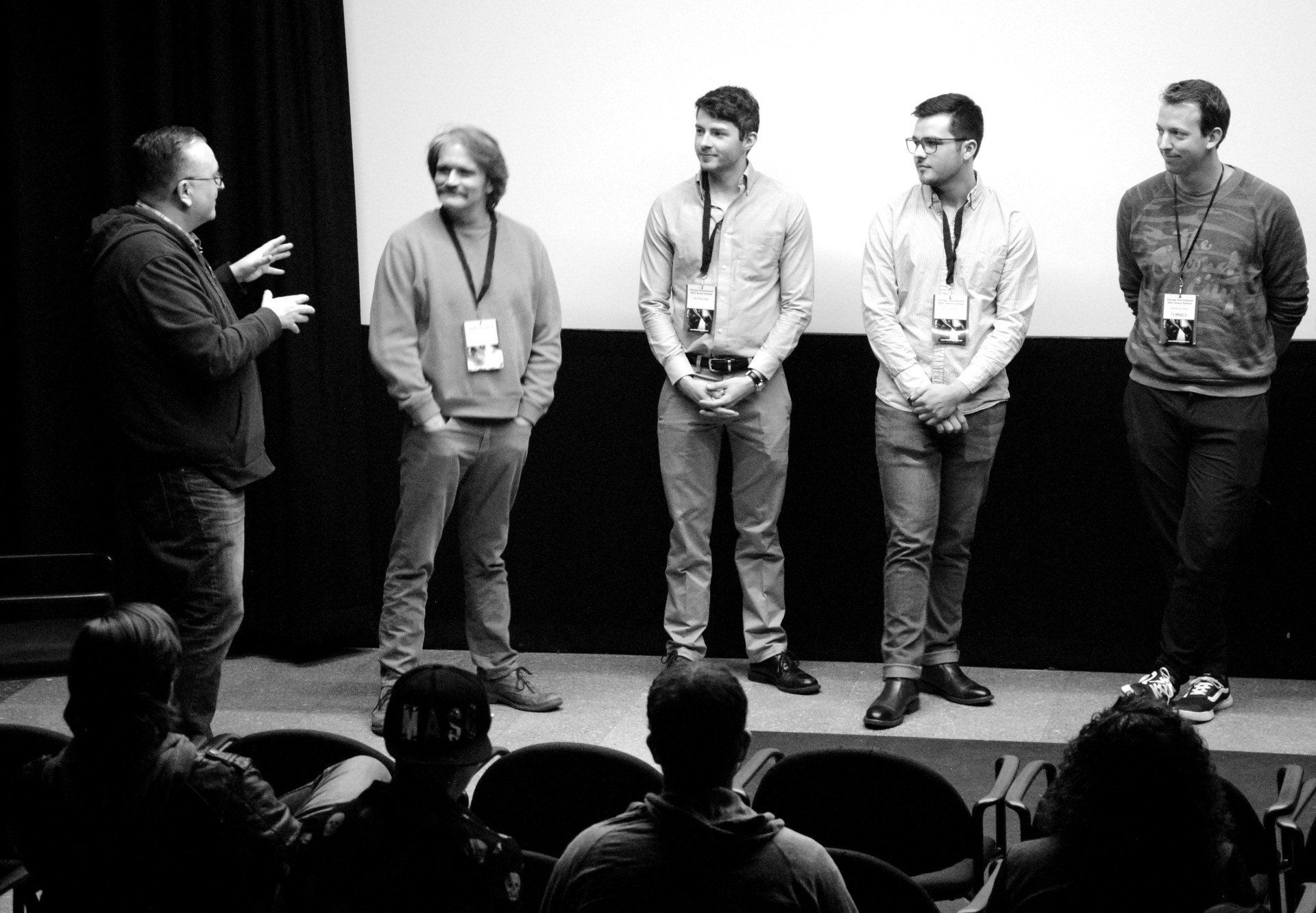 A panel of five men with lanyards on stage; an audience listens. Black and white.