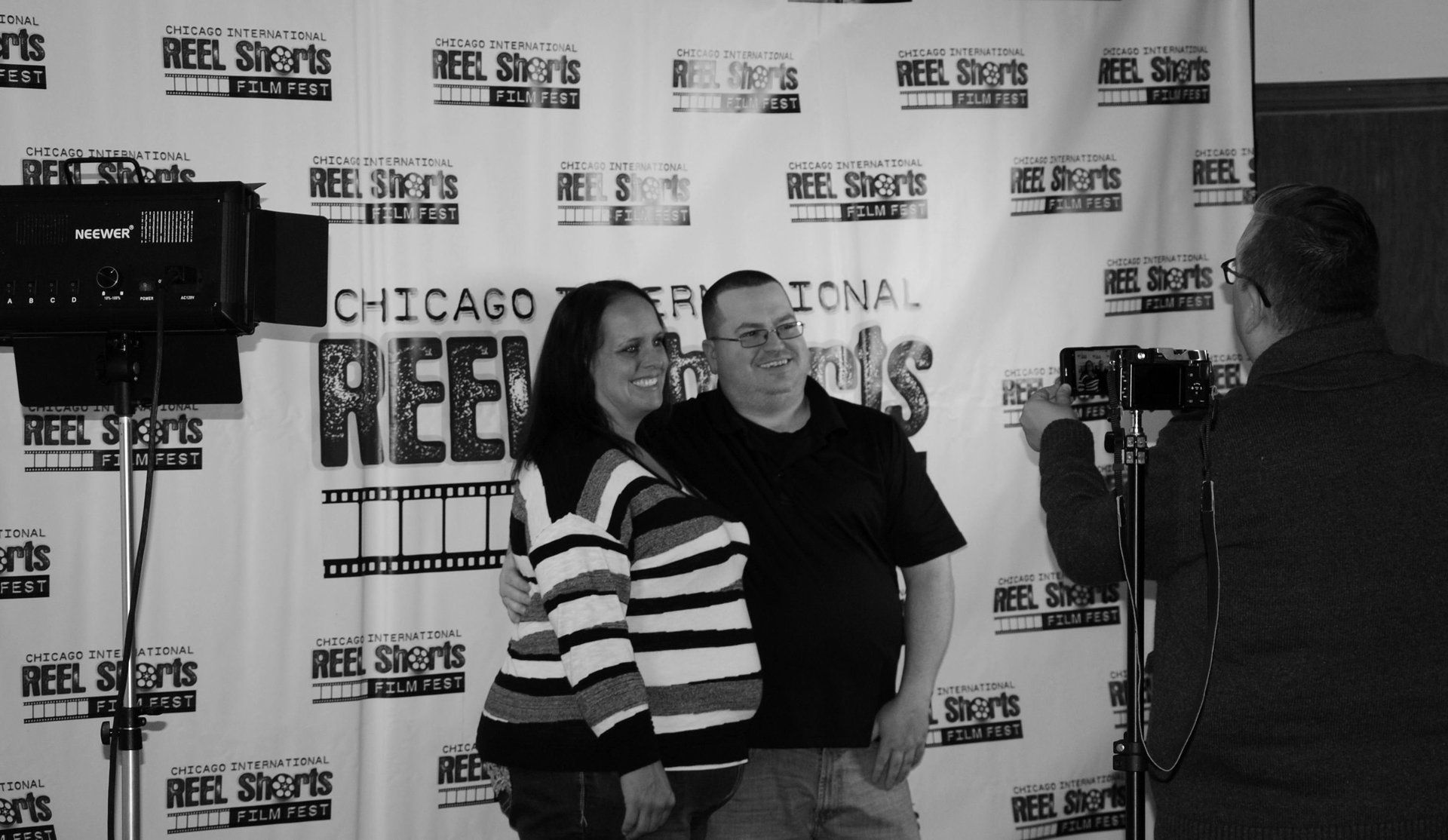 Couple poses for a photo at a film festival with a backdrop that has “REEL Shorts” printed on it.