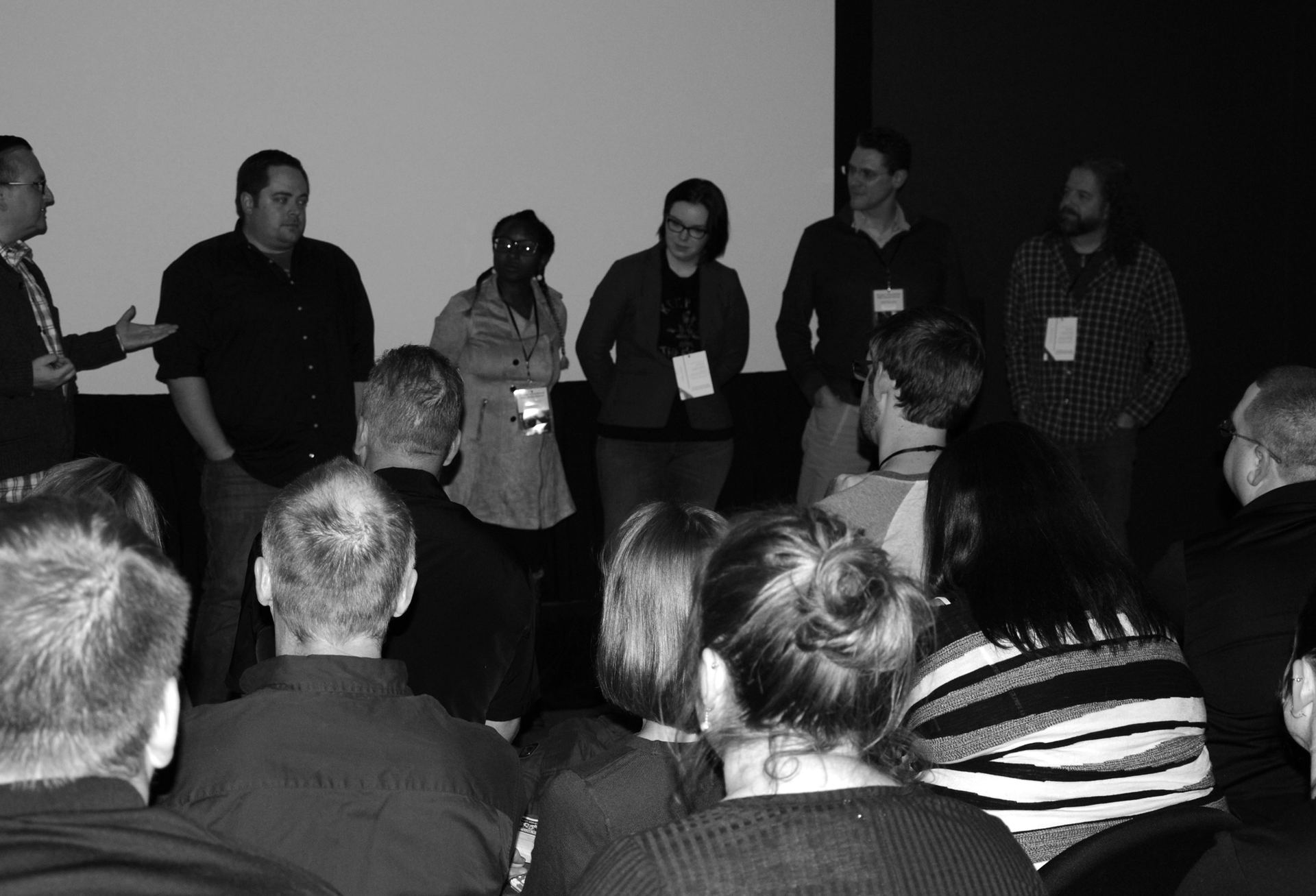 A panel of six people standing on stage, addressing an audience, with a screen behind them.