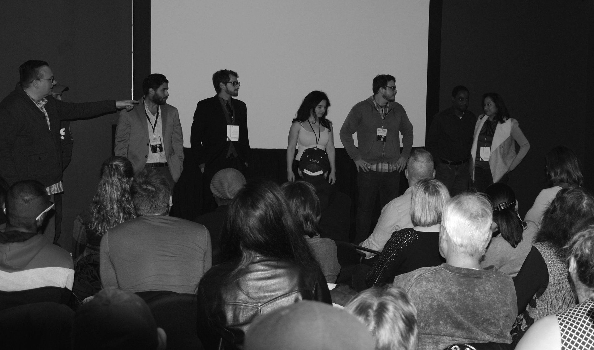A panel of people stand on stage in front of an audience. A man gestures with his arm.