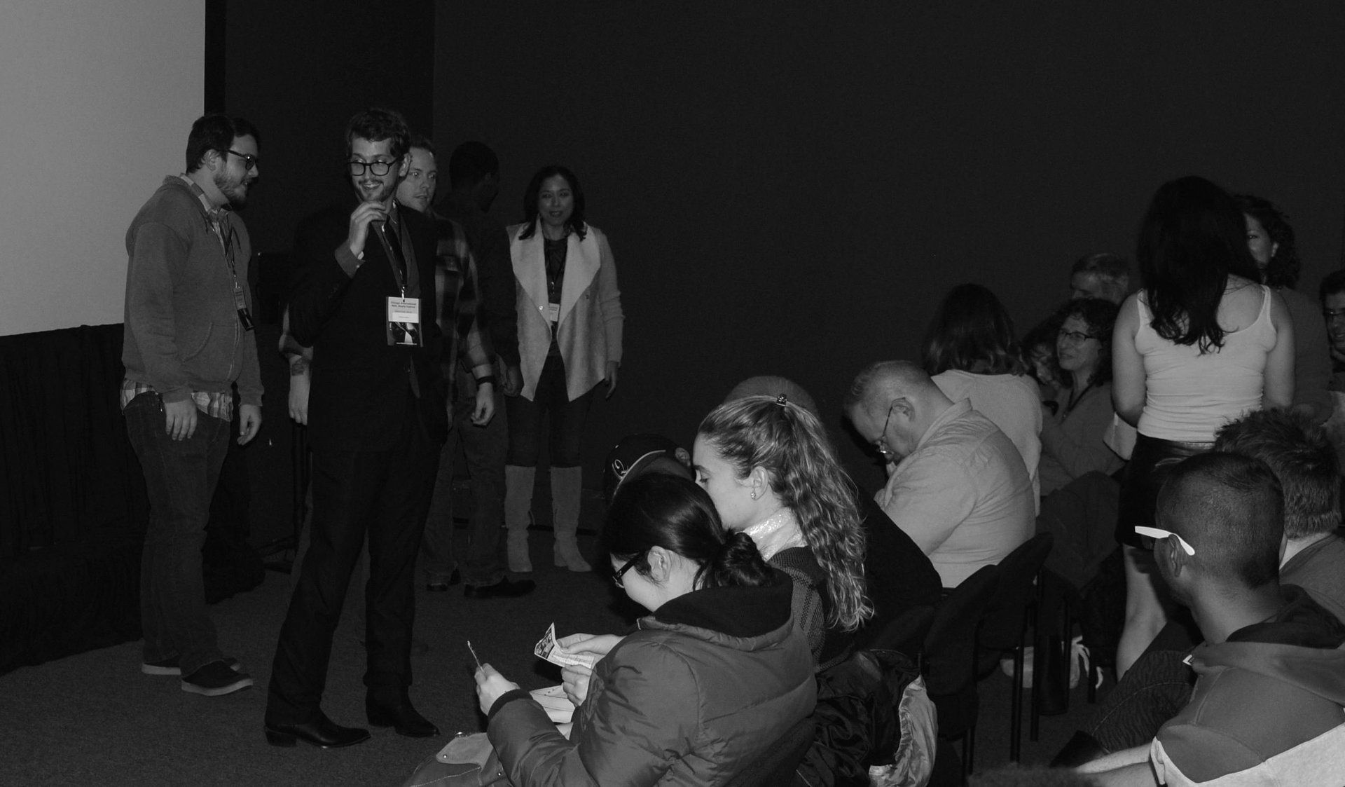 A group of people inside a dark room, some seated while others stand near a presenter.