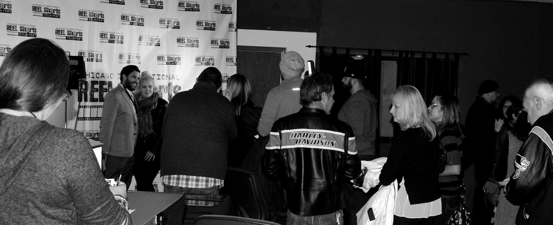 People gathered in a room near a banner. Some are signing things, others are observing. Black and white.