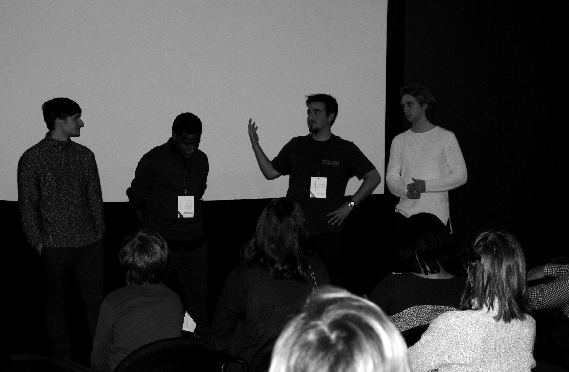Panel of four men speaking on a stage, addressing an audience seated in a dark room.