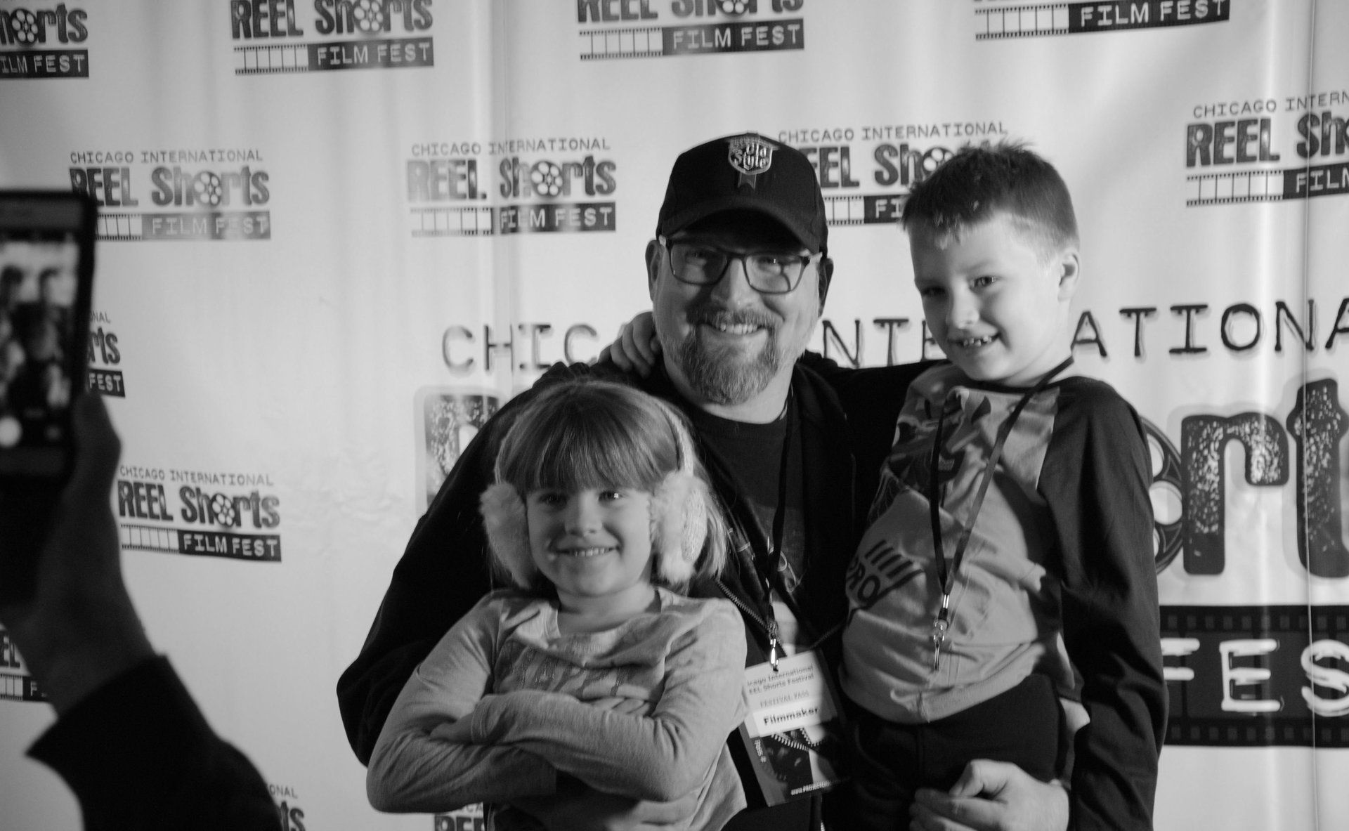 Man and two children smiling at event. Man has glasses and baseball cap. Children wear earmuffs and smile.