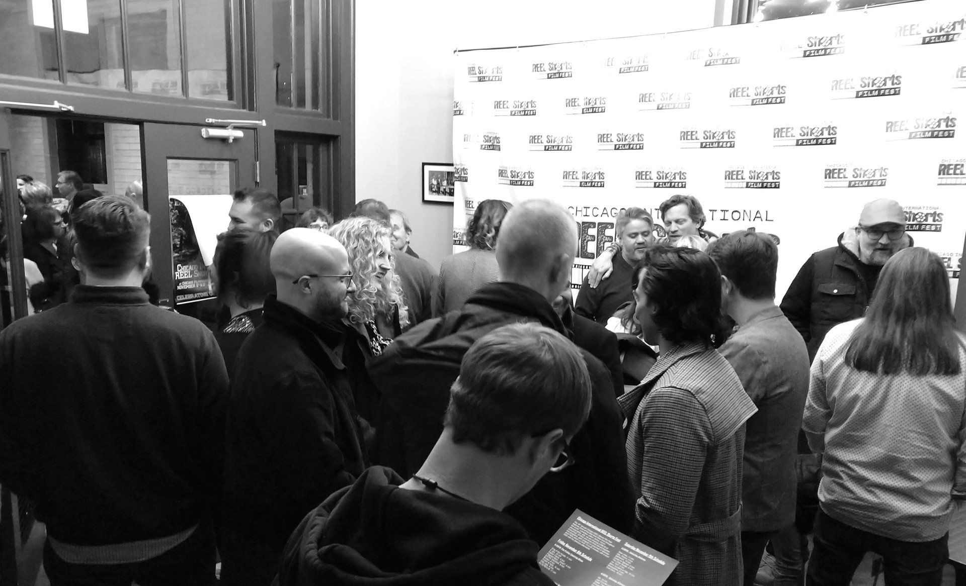 People gathered at an event, possibly a book signing. Several figures interact near a backdrop; some appear to be signing items.