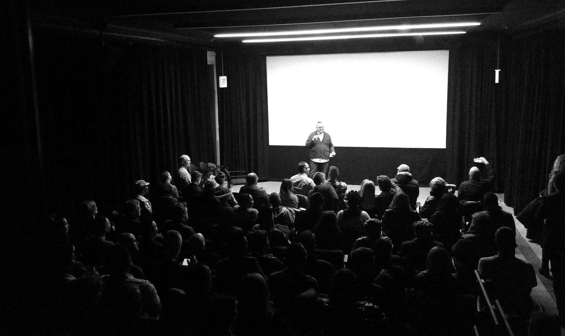 Audience in a dark theater watches a man at a podium in front of a blank screen.