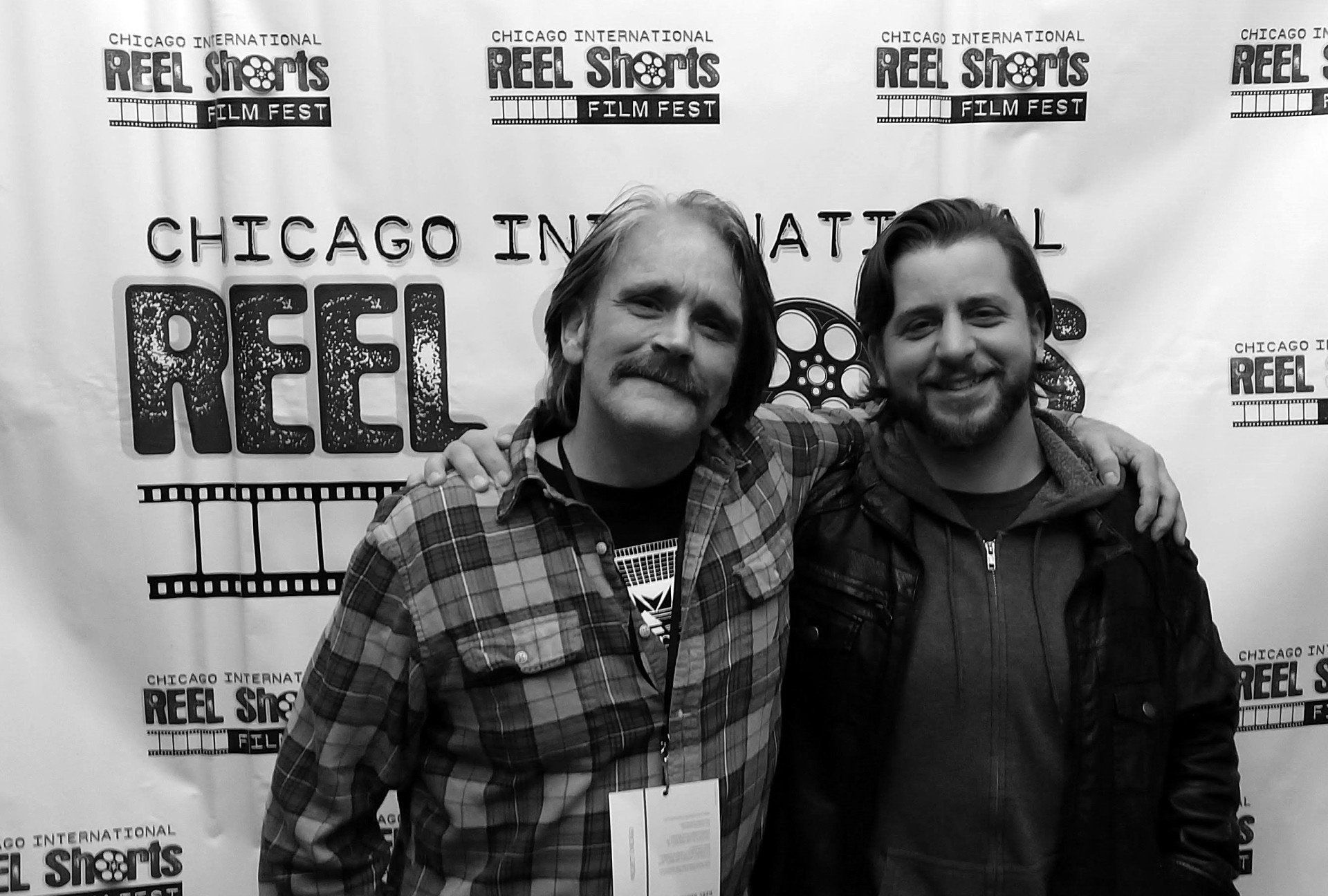 Two men smiling, arms around each other in front of a backdrop for the Chicago International Reel Shorts Film Fest.