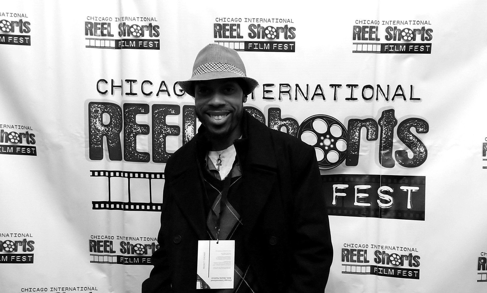 Man smiling at film festival banner. Wearing a fedora, black coat, and a badge.