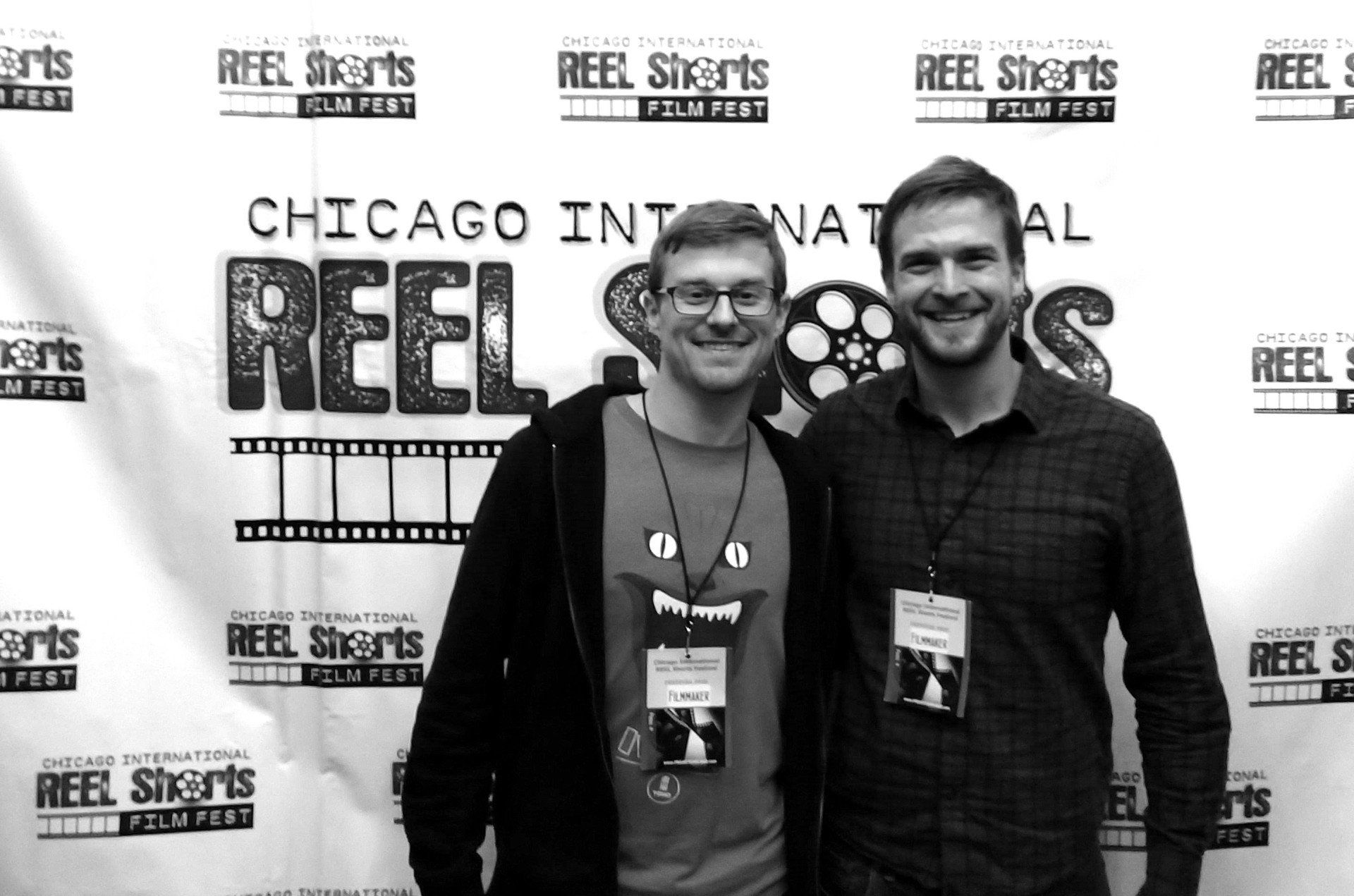 Two men at the Chicago International Reel Shorts Film Fest, posing in front of a banner.