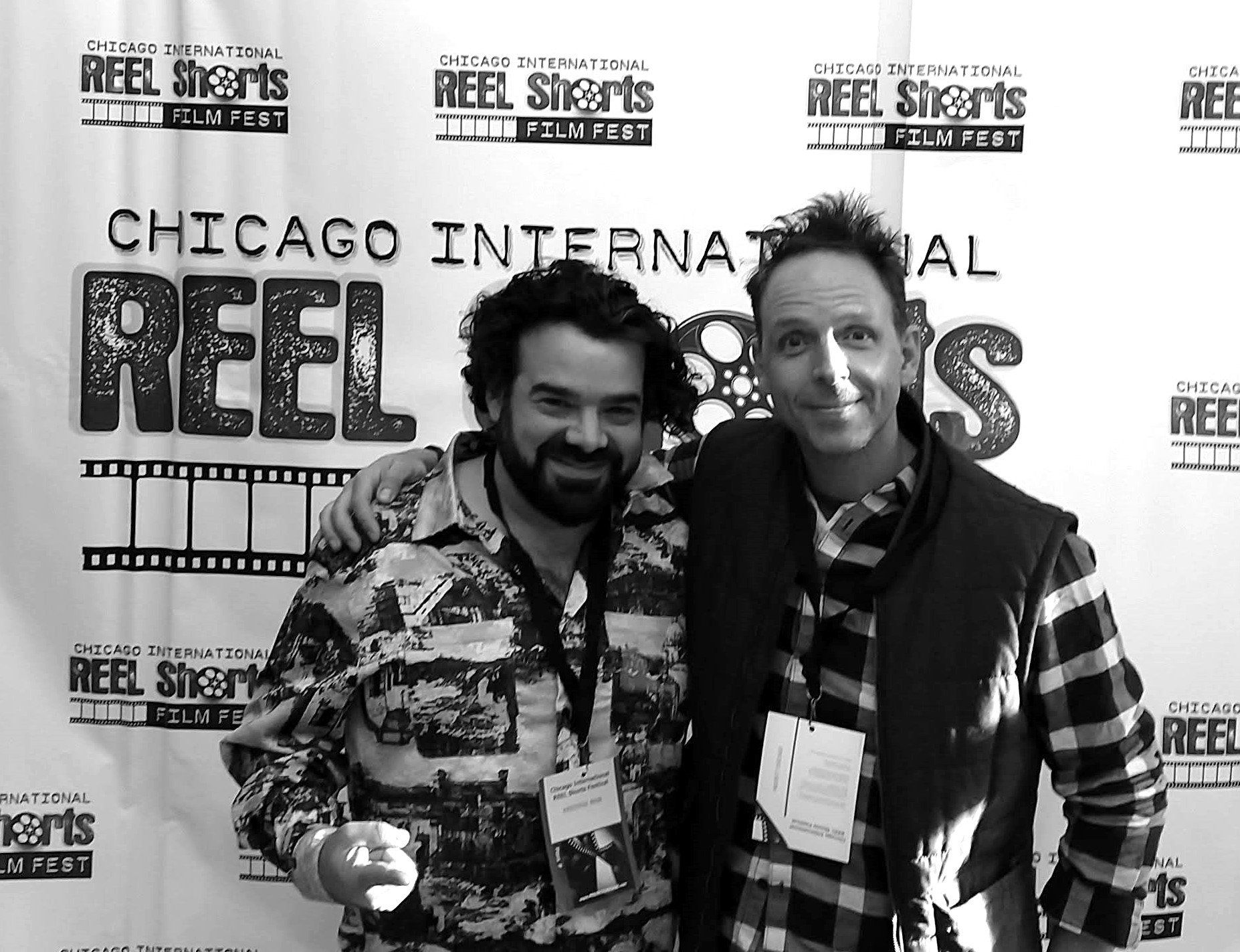 Two men pose in front of a film festival banner. One has an arm around the other, smiling. Black and white.