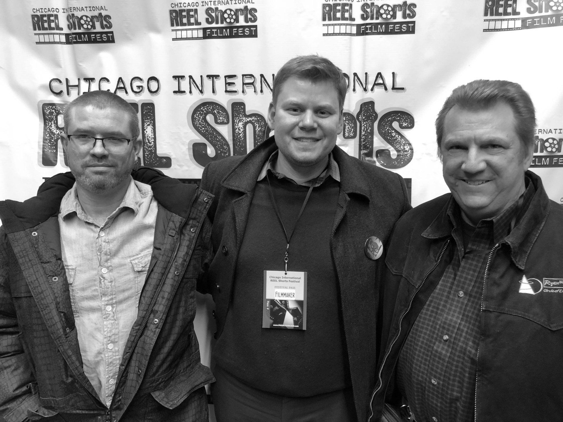 Three men pose at the Chicago International Reel Shorts festival. One wears glasses and a jacket; the others have neutral expressions.
