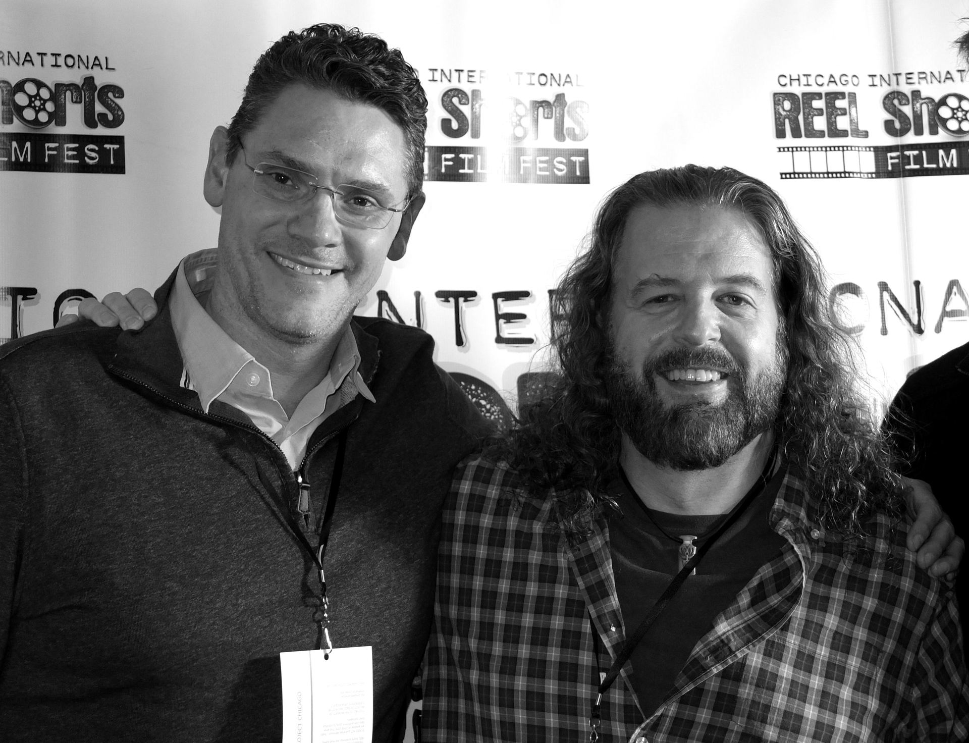 Two men at a film festival posing for a photo, smiling. One wears glasses and the other has long hair and a beard.